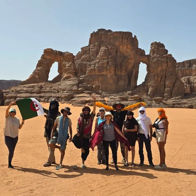 Groupe de voyageurs brandissant le drapeau algérien devant une arche de grès, dans le sable du désert