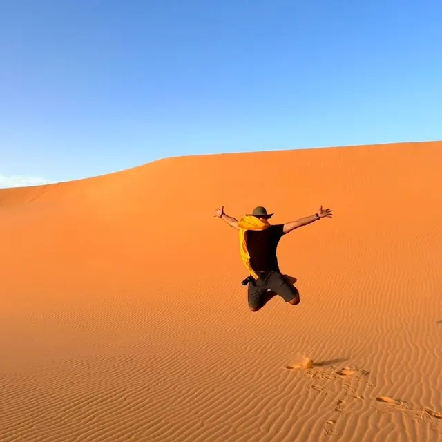 Voyageur sautant de joie sur une dune orangée, sous un ciel bleu