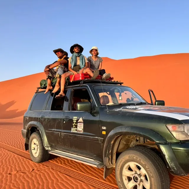 Trois voyageurs assis sur le toit d'un 4×4, devant une grande dune rouge