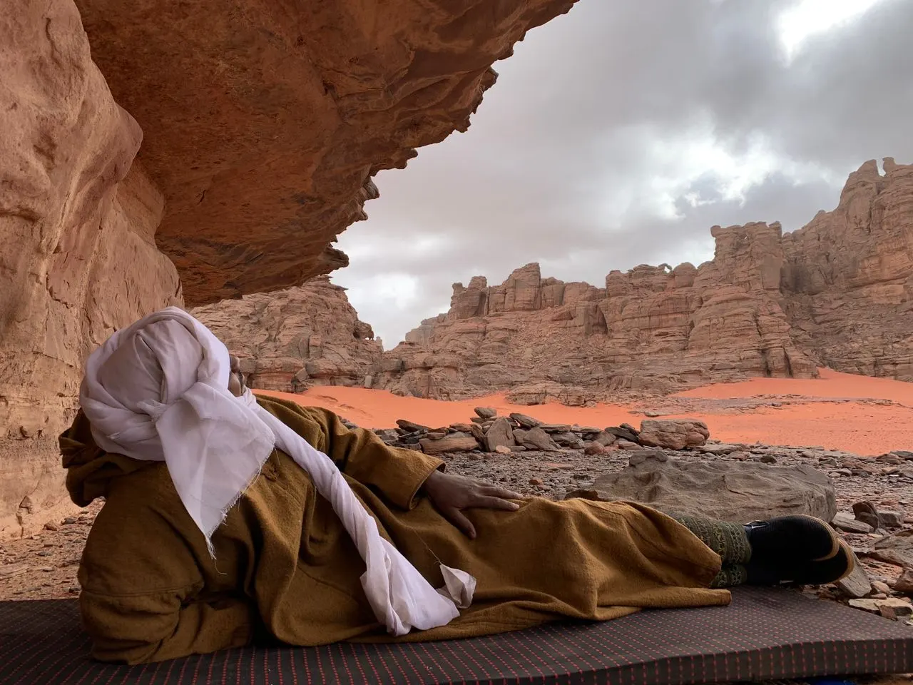 Guide touareg au repos sous un surplomb rocheux, face aux falaises et au sable rouge